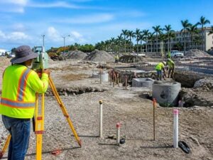 Construction site with a surveyor in a neon vest using a level, while workers install foundations and pipes in the background.